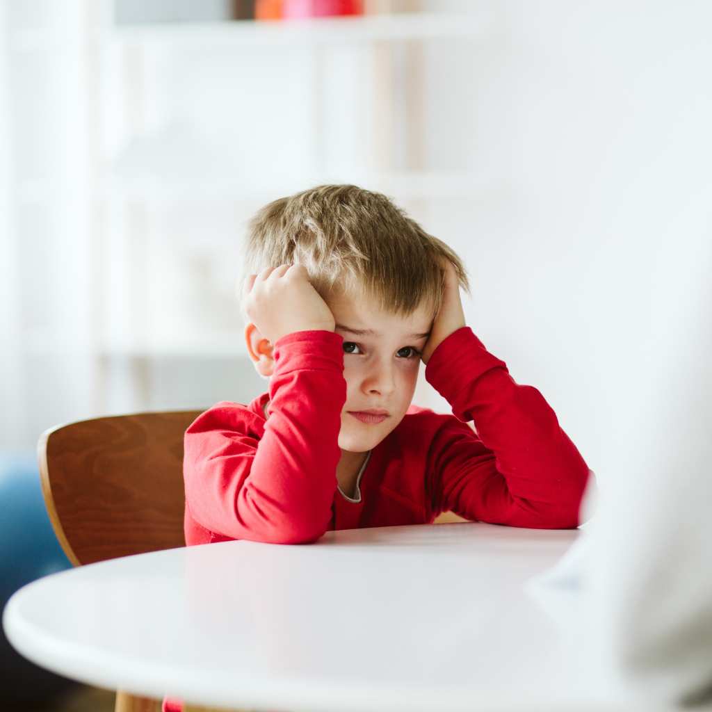 A young child on the spectrum sitting at a table with their hands on their head, appearing stressed or overwhelmed
