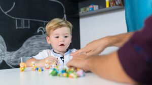 Toddler boy engages with small toy figures on a table while a therapist guides him during an early intervention for autism in a clinic with a chalkboard behind them